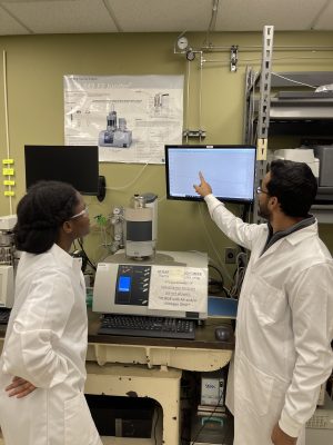 two research students in front of a monitor in white lab coats discussing clean energy